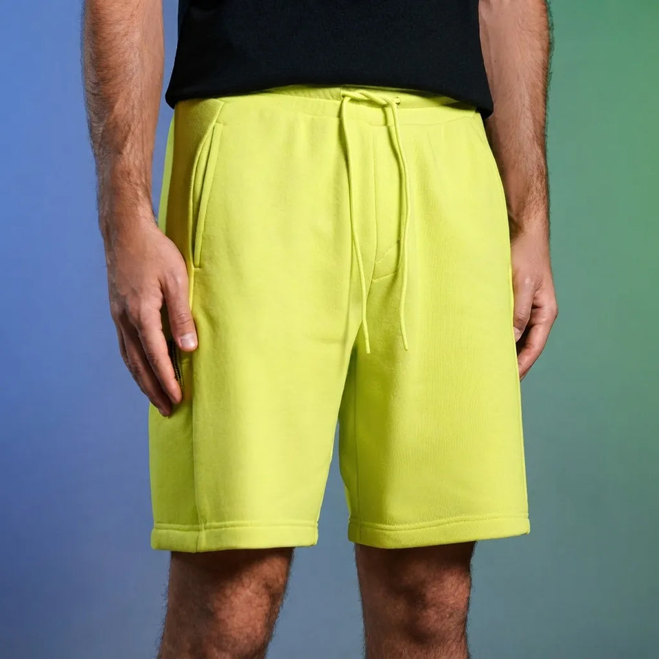 A person wearing bright neon yellow drawstring shorts and a black top, standing against a blue-to-green gradient studio background.