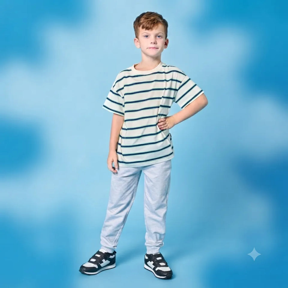 A young boy stands against a blue studio backdrop wearing a short-sleeve striped t-shirt, light gray sweatpants, and black-and-white sneakers.