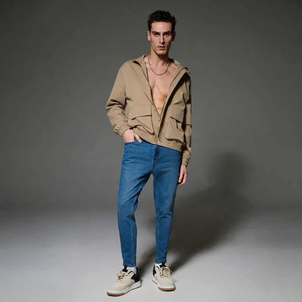 A male model stands against a dark gray studio background wearing a beige zip-up hooded jacket, blue jeans, and off-white sneakers.