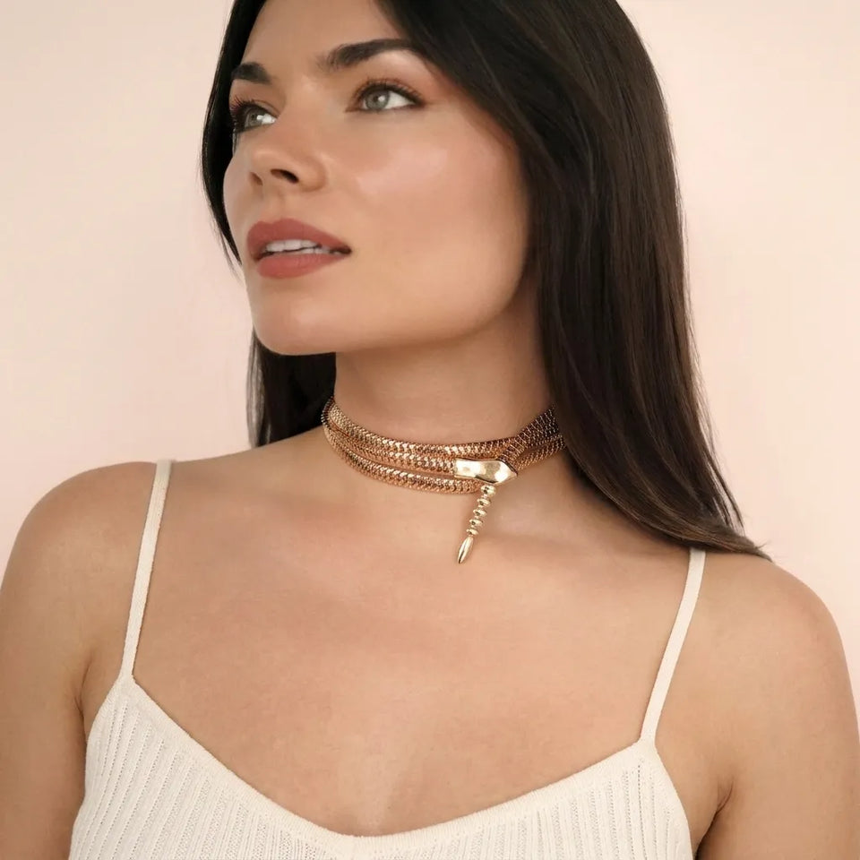 A woman with long dark hair models a multi-layered rose gold choker necklace with a small dangling charm, wearing a cream-colored ribbed camisole against a soft beige background.