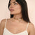 A woman with long dark hair models a multi-layered rose gold choker necklace with a small dangling charm, wearing a cream-colored ribbed camisole against a soft beige background.