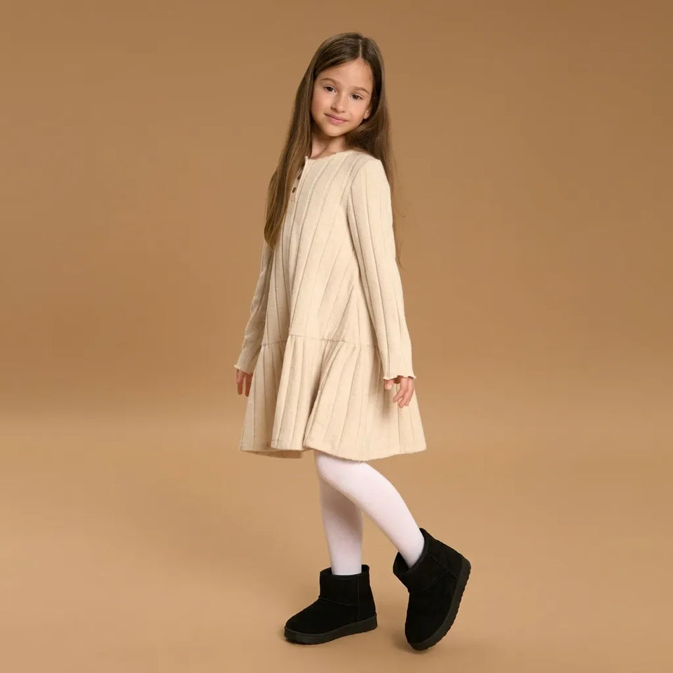 A young girl with long brown hair stands in a studio wearing a cream-colored ribbed knit long-sleeve dress, white tights, and black boots against a solid tan background.