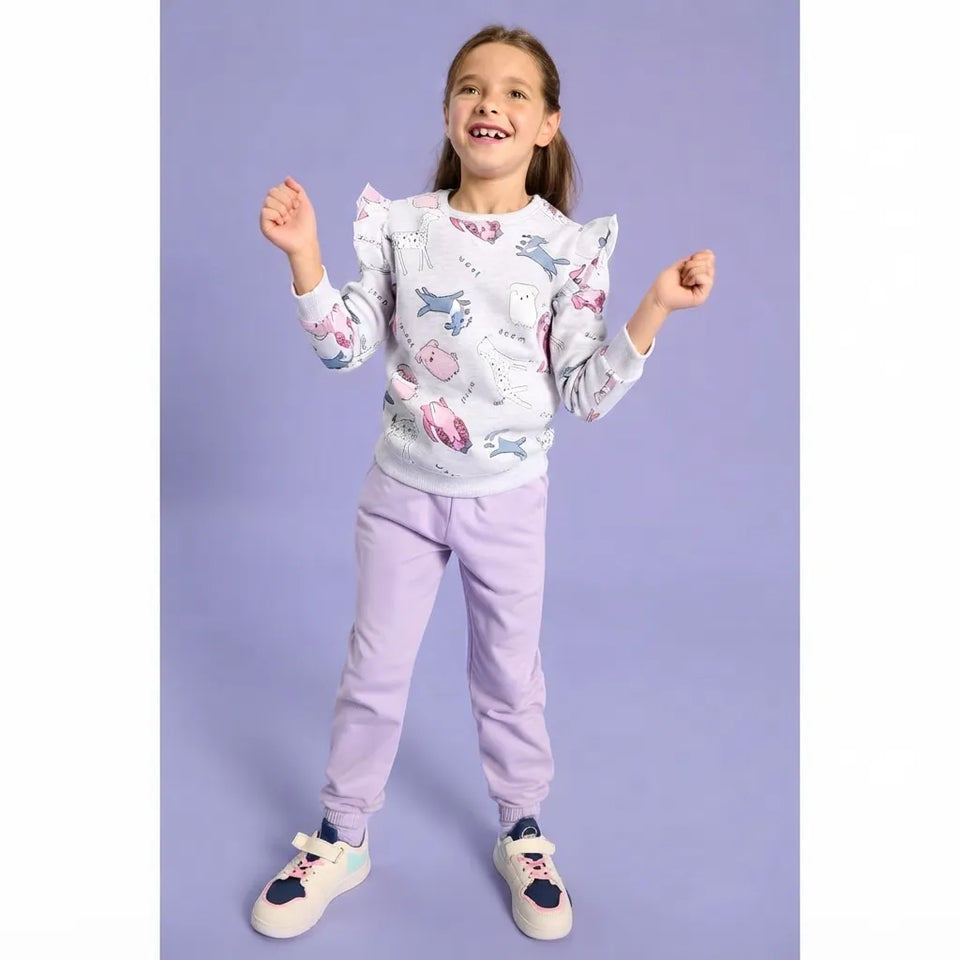 A young girl smiling and posing in a white long-sleeve sweatshirt with a colorful dinosaur print, paired with light purple jogger pants and white sneakers, set against a solid lavender studio background.