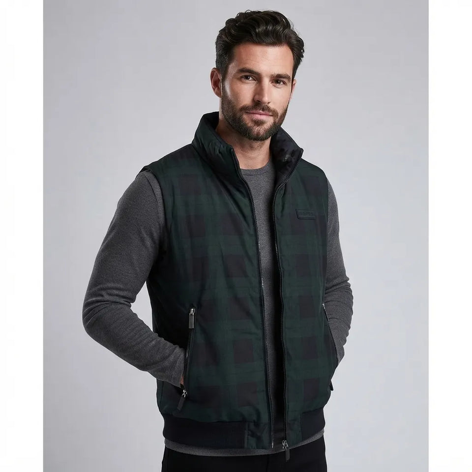 A man wearing a dark green and black plaid zip-up vest over a dark gray long-sleeve shirt, photographed against a neutral gray studio background.