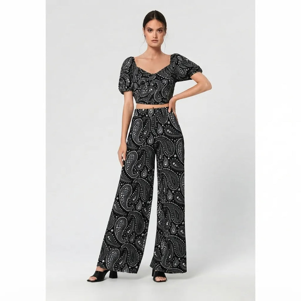 A woman modeling a black two-piece outfit with a white paisley pattern, featuring a short-sleeve crop top with puff sleeves and matching wide-leg pants, set against a plain light gray studio background.