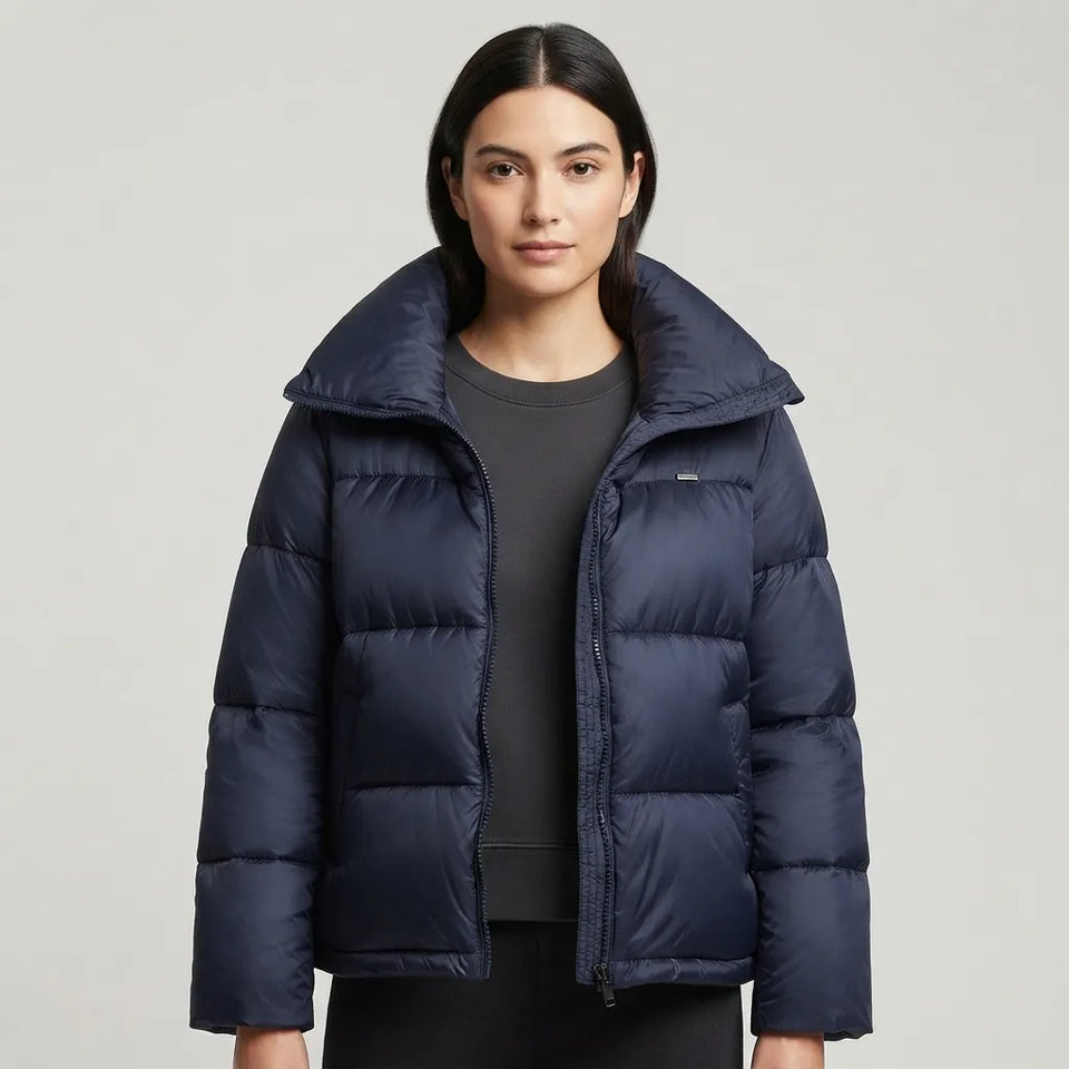 A woman modeling a navy blue puffer jacket with a high collar and horizontal quilting, worn over a dark gray top against a light gray studio background.