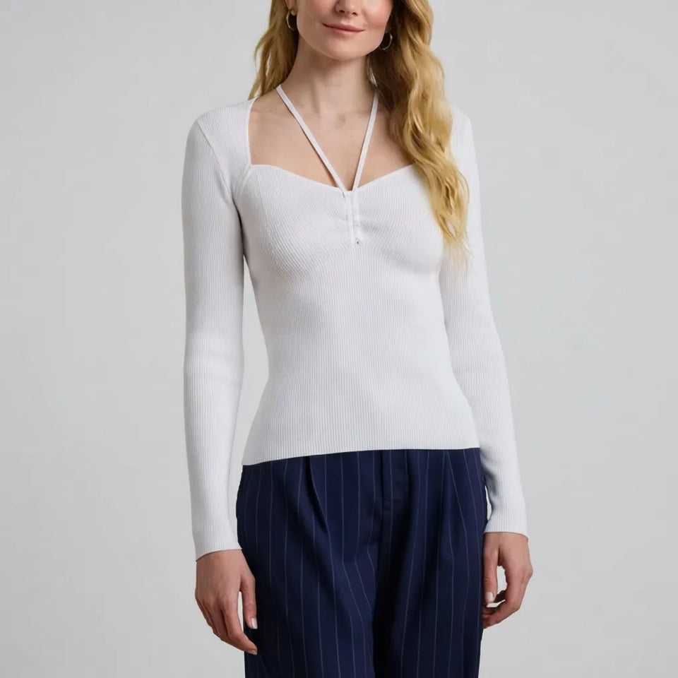 A woman wearing a fitted white ribbed knit long-sleeve top with a square neckline and double crossover front straps, paired with navy pinstriped trousers, against a light gray studio background.