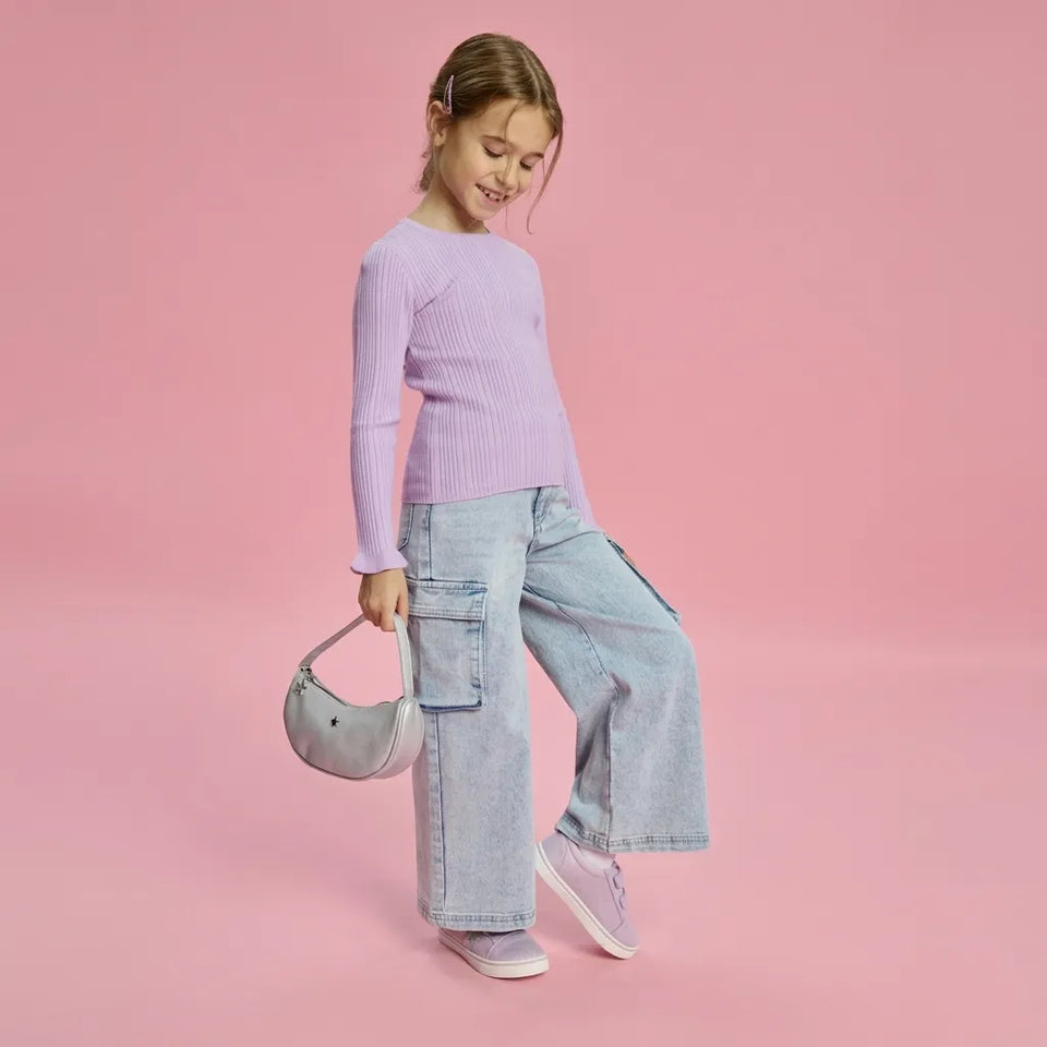 A young girl modeling a light purple ribbed long-sleeve top, light-wash wide-leg denim cargo pants, and lavender sneakers, holding a small gray crescent-shaped handbag against a solid pink background.