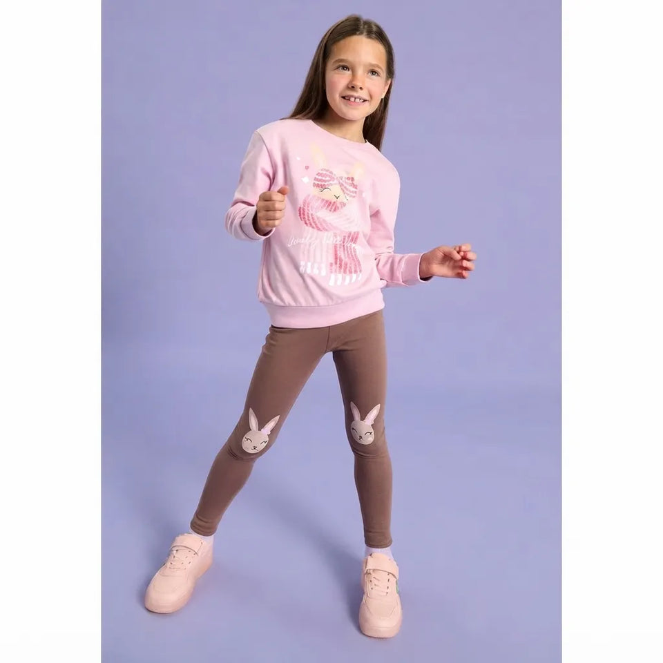 A young girl modeling a pink long-sleeve sweatshirt with a colorful cupcake graphic, paired with brown leggings featuring bunny patches on the knees, and light pink sneakers, set against a solid lavender studio background.