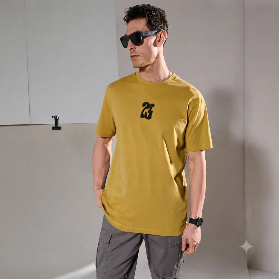 A man wearing a mustard yellow t-shirt with a black graphic logo on the chest, styled with gray cargo pants, black sunglasses, and a black watch, standing against a minimalist gray wall.