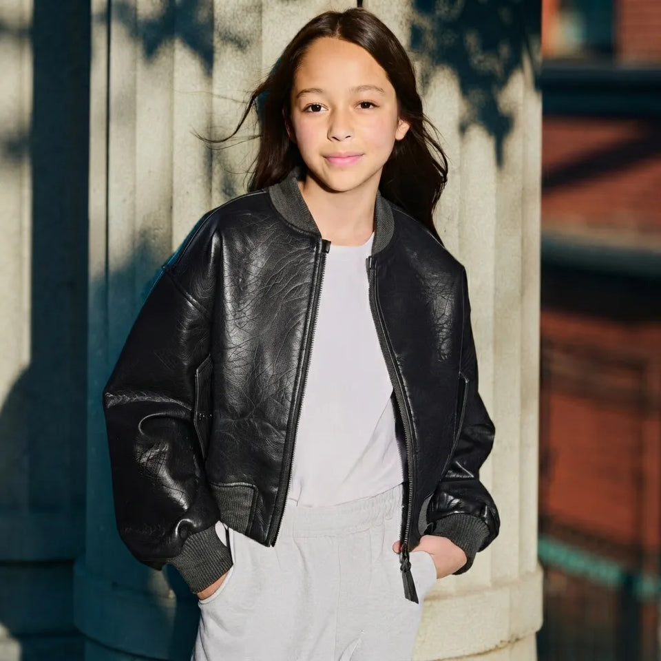 A young girl wearing a black faux-leather bomber jacket over a white t-shirt and light gray sweatpants, standing outdoors in front of a stone column.