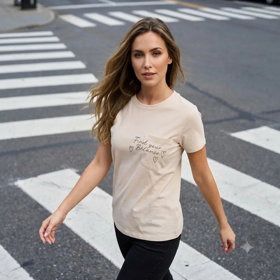 A woman walking on a city crosswalk wearing a light beige crewneck t-shirt with a small chest pocket and black pants. The t-shirt features a cursive graphic that reads 'Find your Balance' with small hearts.