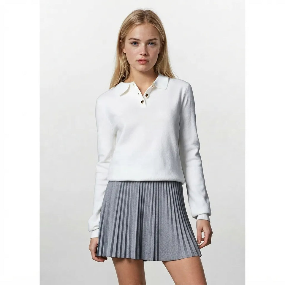 A blonde model wearing a white long-sleeve polo sweater and a gray pleated mini skirt, standing against a light gray studio background.