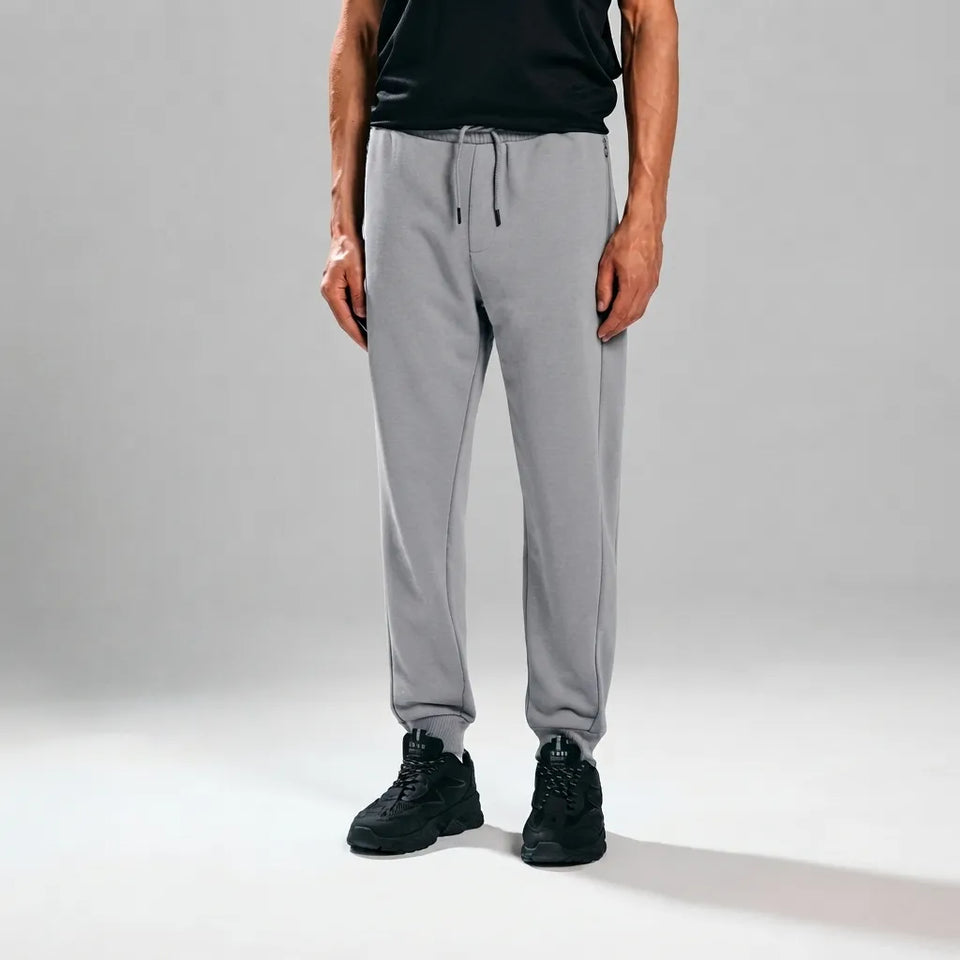 A person wearing light gray jogger sweatpants with a drawstring waist, paired with a black shirt and black sneakers, standing against a light gray studio background.