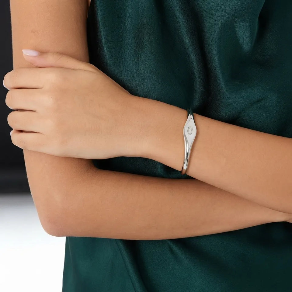 A close-up of a person wearing a polished silver cuff bracelet with a small engraved symbol, paired with a dark green satin top.