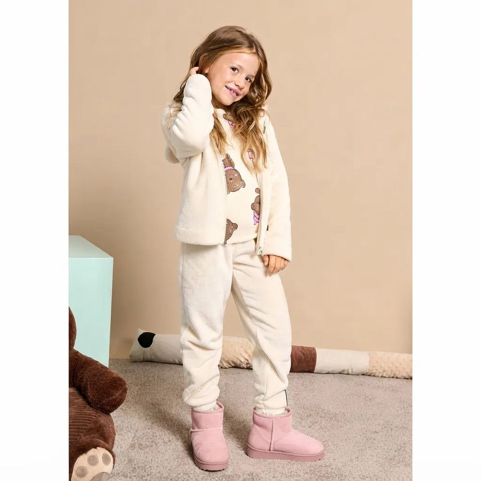 A young girl stands in a room wearing a cream-colored fleece pajama set with a teddy bear print, matching cream sweatpants, and pink slip-on boots.
