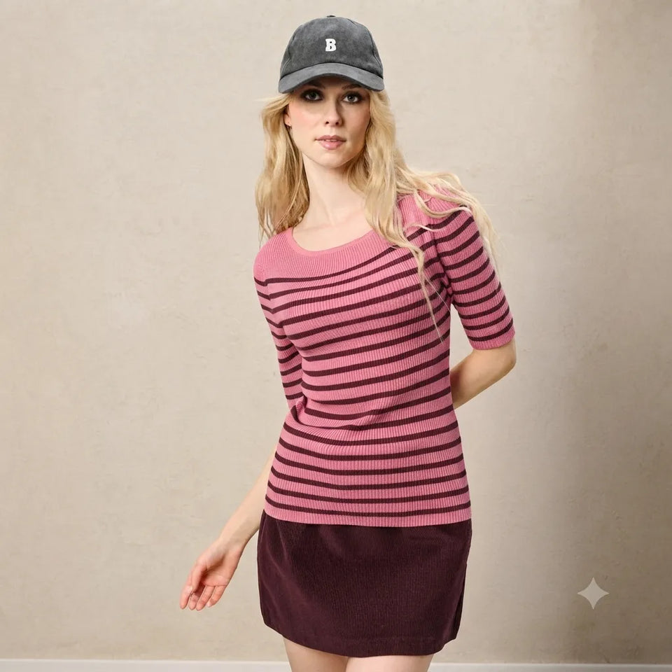 A woman wearing a pink and dark red striped short-sleeve knit top, a dark plum skirt, and a gray baseball cap with a white letter 'B' on the front, standing against a neutral beige wall.