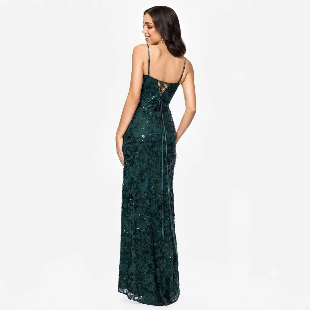 A woman modeling a floor-length dark green sequined gown from the back, featuring thin straps, a lace-embroidered bodice, and a lace-trimmed back slit, set against a plain white studio background.