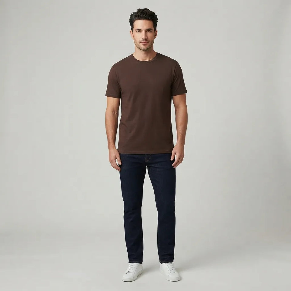 A man modeling a dark brown short-sleeve crewneck t-shirt, paired with dark navy jeans and white sneakers, standing against a light gray studio background.
