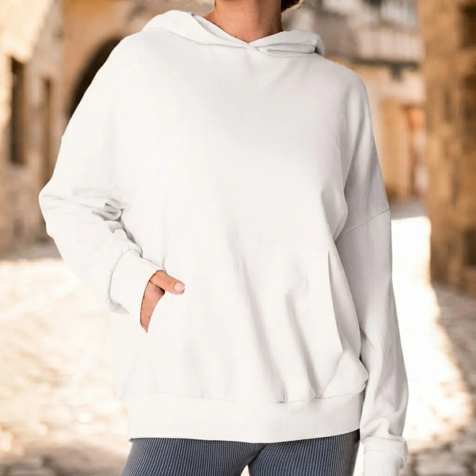 A person wearing an oversized white pullover hoodie with a kangaroo pocket, standing in a sunlit outdoor alley with blurred stone buildings in the background.