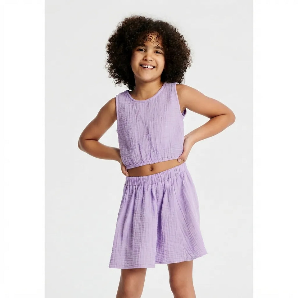 A young girl with curly dark hair wearing a light purple two-piece set consisting of a sleeveless cropped top and a matching flared skirt, posed against a light gray studio background.