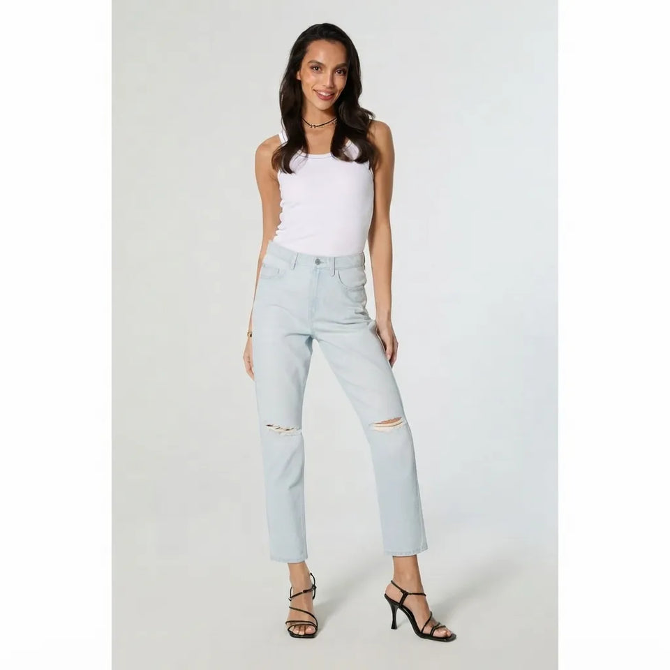 A woman modeling light-wash distressed denim jeans with a white sleeveless top and black strappy heels, standing against a light gray studio background.