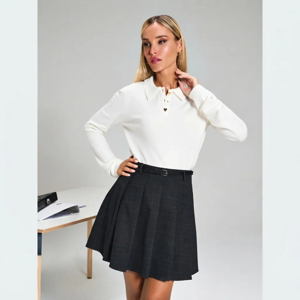A woman modeling a white long-sleeve polo shirt and a black pleated mini skirt with a subtle windowpane pattern, standing in a studio setting.