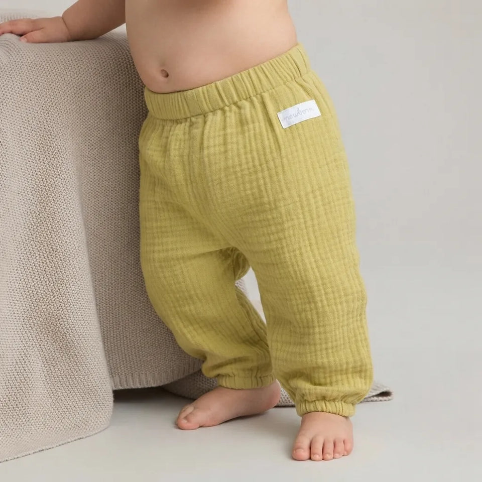 A baby wearing textured olive green gauze pants, standing barefoot next to a beige knit blanket.