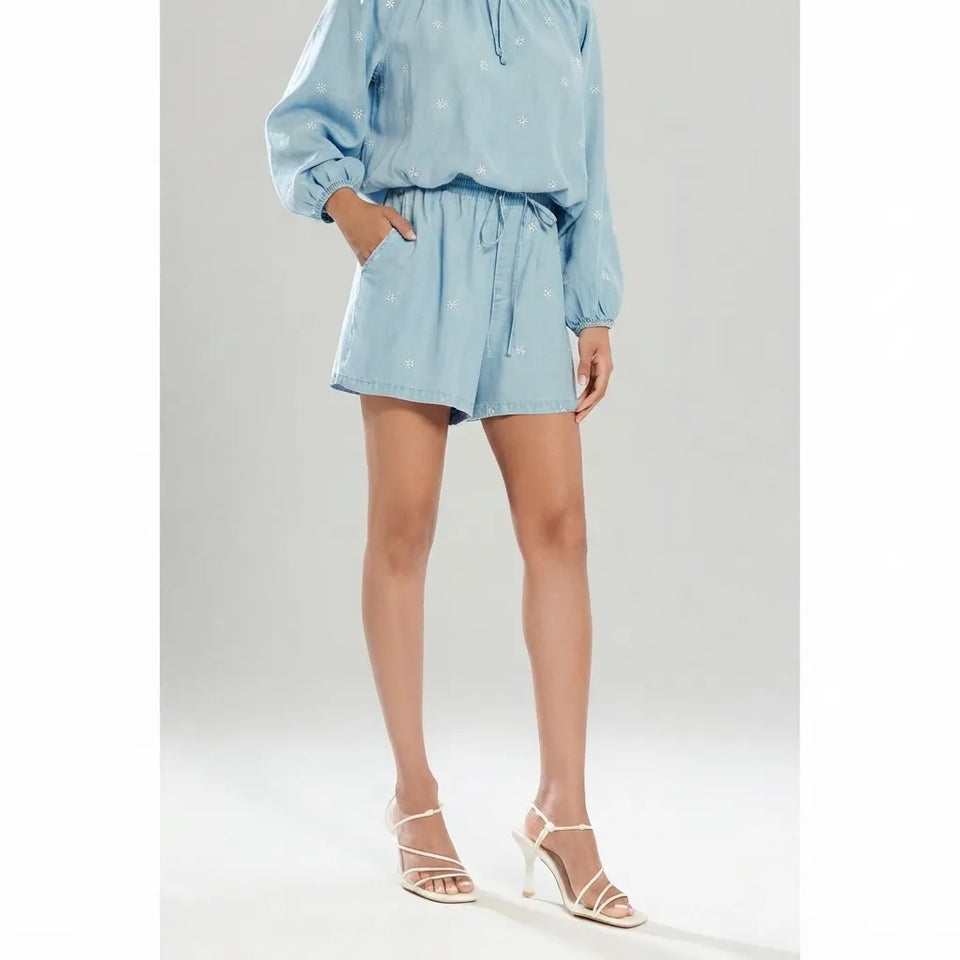 A model wearing a light blue two-piece set consisting of a long-sleeve top and matching shorts with a subtle white pattern, paired with white strappy high-heeled sandals against a light gray studio background.