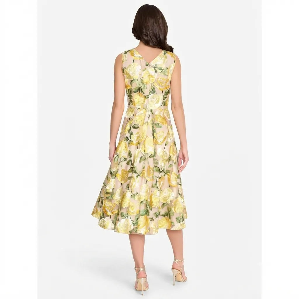 A woman viewed from the back wearing a sleeveless midi dress with a yellow and green floral print, paired with beige high-heeled sandals, against a light gray studio background.