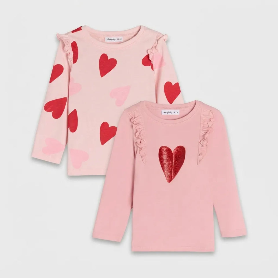 Two pink long-sleeve children's tops with ruffle details, displayed on a light gray background. The top on the left has scattered red and pink hearts, while the top on the right features a large red glitter heart on the chest.