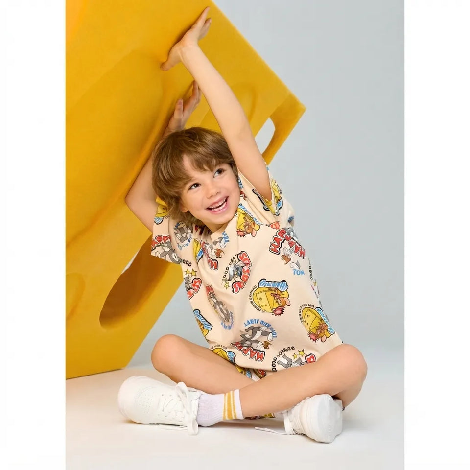 A young boy sitting on the floor, smiling and stretching his arms up to hold a large yellow geometric prop. He is wearing a beige short-sleeve t-shirt with a colorful cartoon graphic print, white socks, and white sneakers.