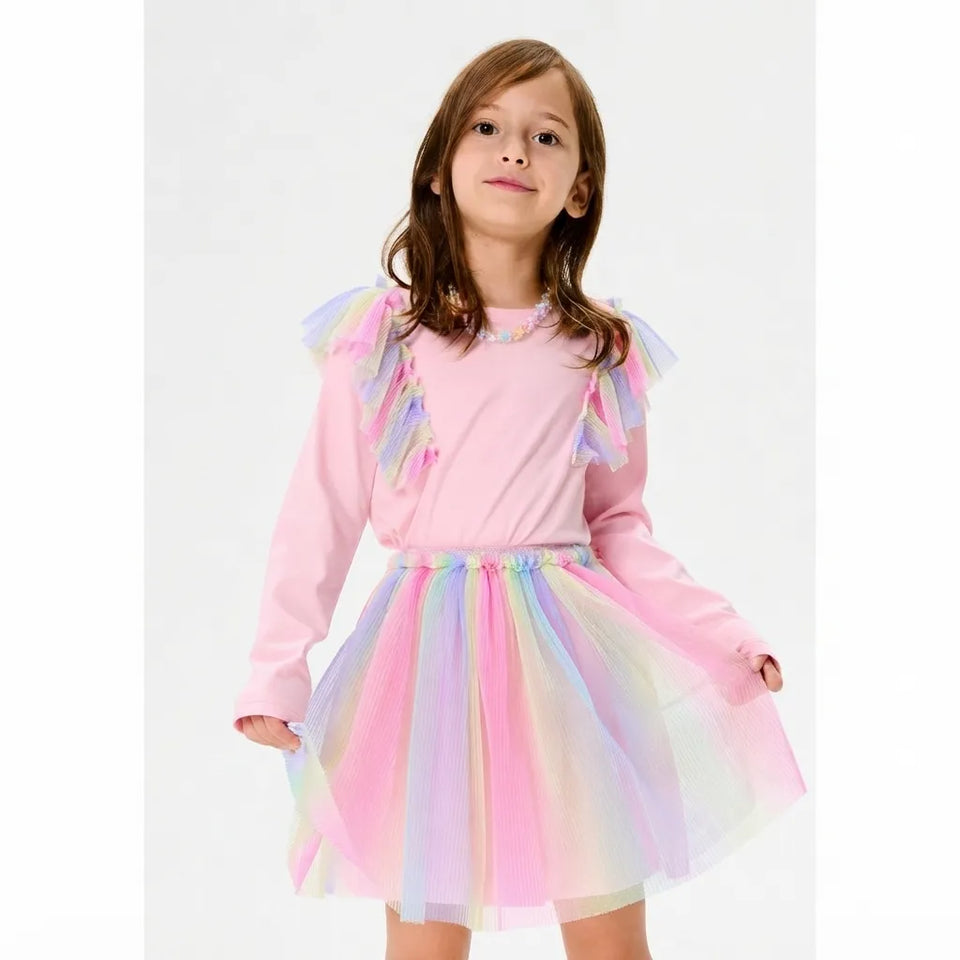 A young girl wearing a pink long-sleeve dress with a pastel rainbow tulle tutu skirt and matching ruffled shoulder details, photographed against a plain light gray studio background.