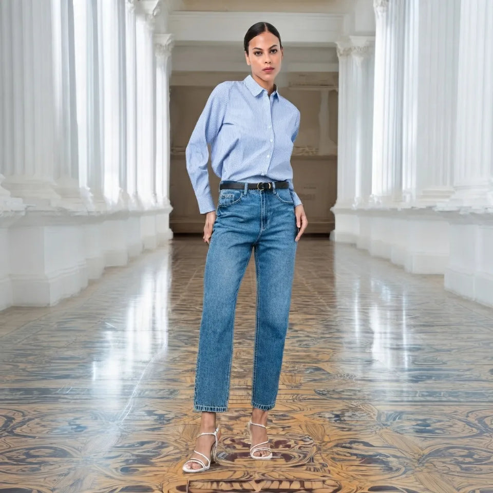 A woman stands in a grand white-columned hallway with a patterned floor, wearing a light blue button-up shirt, blue jeans, a black belt, and white heeled sandals.