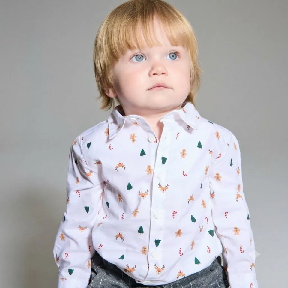 A toddler with blonde hair and blue eyes wearing a white long-sleeve button-up shirt with a festive pattern of green pine trees, brown reindeer, and candy canes, paired with gray pants, photographed against a neutral gray background.