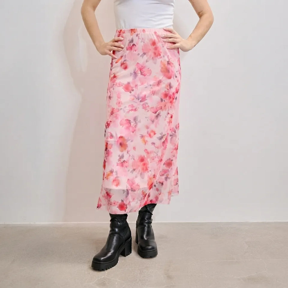 A model wearing a pink floral midi skirt with a white top and black platform boots, standing against a plain light gray wall.