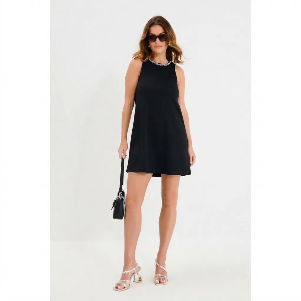 A woman modeling a sleeveless black mini dress with a scalloped neckline, styled with clear rhinestone-embellished heeled sandals, sunglasses, and a small black handbag against a plain white studio background.