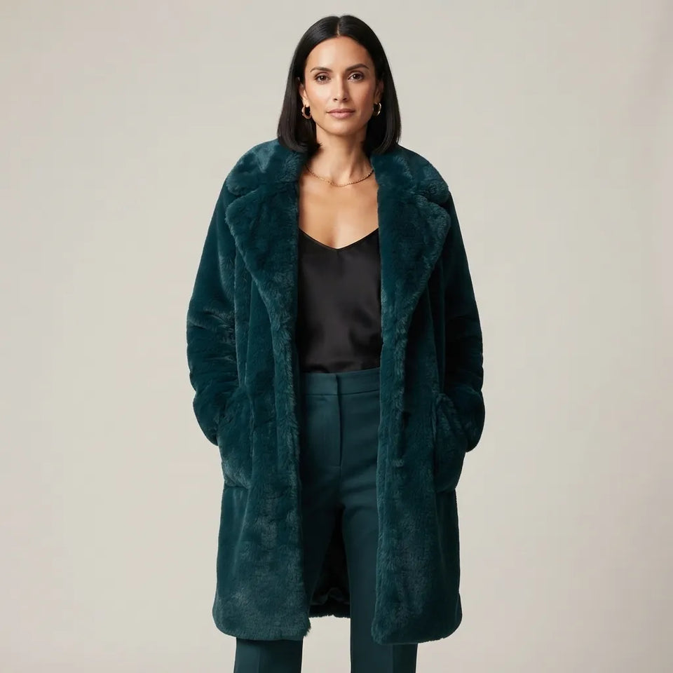 A woman modeling a long, plush dark teal faux fur coat over a black satin top and matching teal trousers, photographed against a neutral light gray studio background.