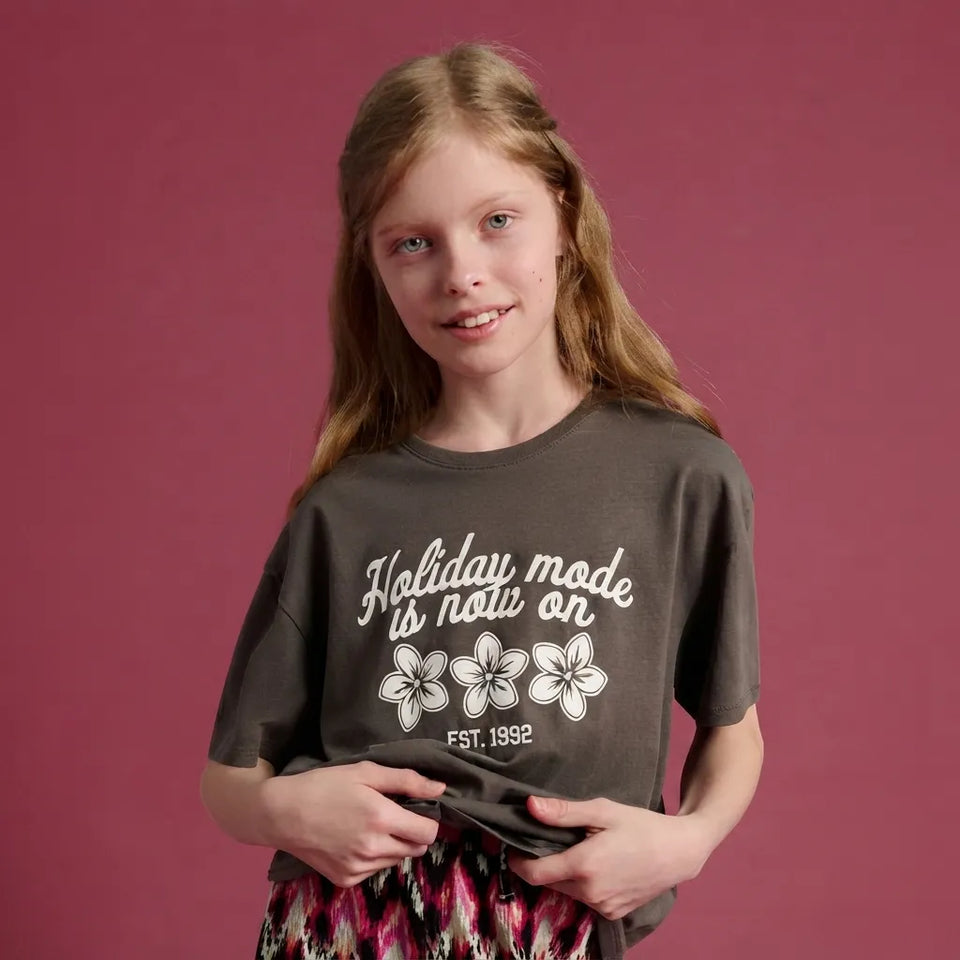 A young girl with long light brown hair stands against a solid dusty rose background wearing a dark brown graphic t-shirt with white text reading 'Holiday mode is now on' and three white flowers, paired with a pink and black patterned skirt.