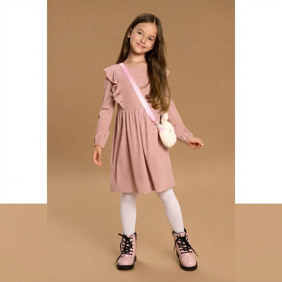 A young girl with long brown hair stands against a solid tan studio background wearing a dusty pink long-sleeve dress with ruffle details, white tights, pink lace-up boots, and a small white bunny-shaped crossbody bag.