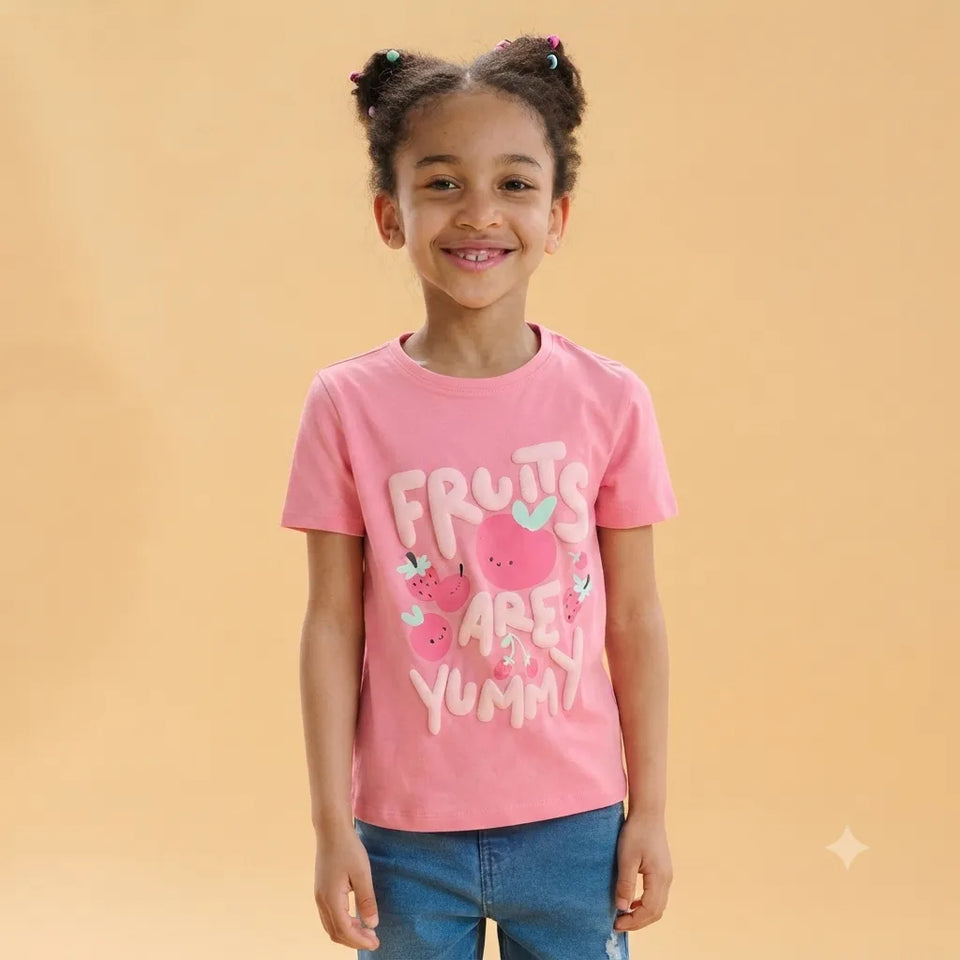 A young girl with her hair in two buns is smiling and wearing a pink short-sleeve t-shirt with a 'FRUITS ARE YUMMY' graphic, paired with blue jeans, against a solid beige background.