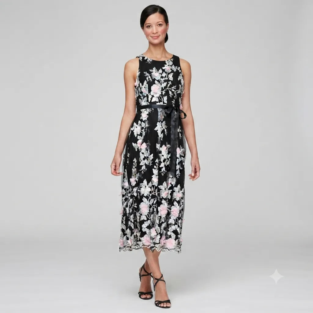 A woman modeling a sleeveless midi dress with a black base and a white and pink floral pattern, featuring a matching black waist tie, against a light gray studio background.