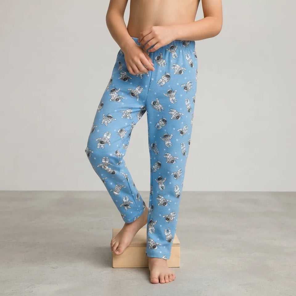 A child wearing light blue pajama pants with a white and gray astronaut print, standing barefoot on a small wooden block against a neutral studio background.