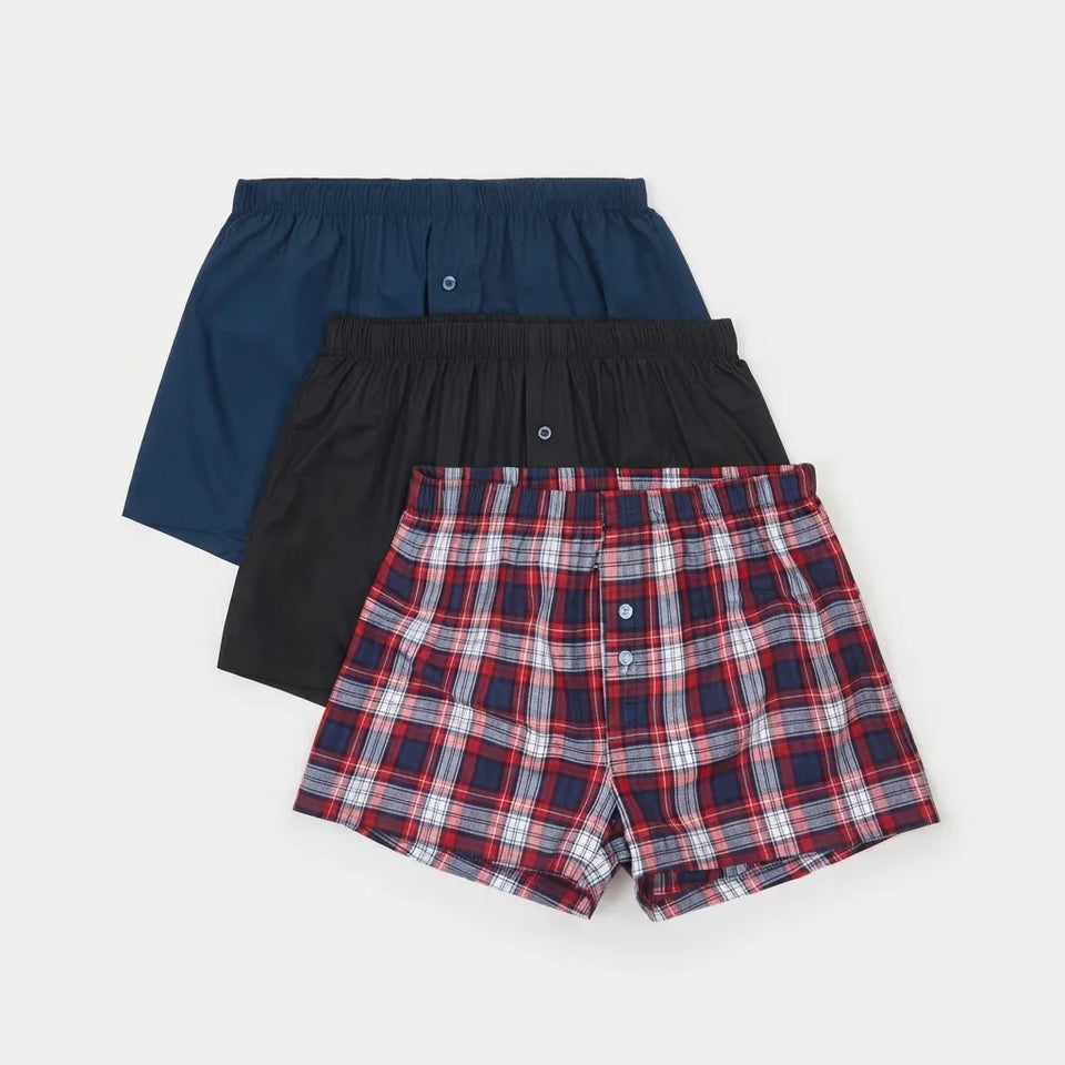 Three pairs of men's boxer briefs arranged in a stack on a light gray background: one navy blue, one black, and one red, navy, white, and gray plaid.