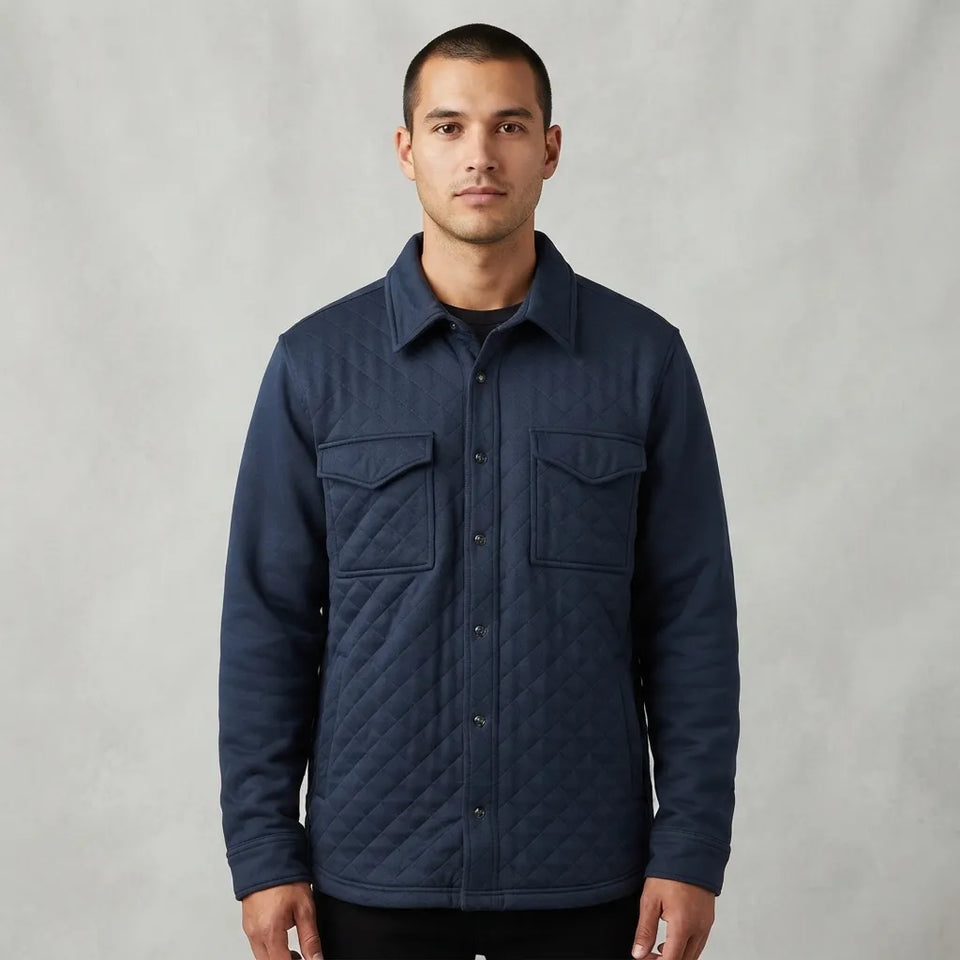 A man wearing a navy blue quilted button-up overshirt jacket, standing against a neutral gray studio backdrop.
