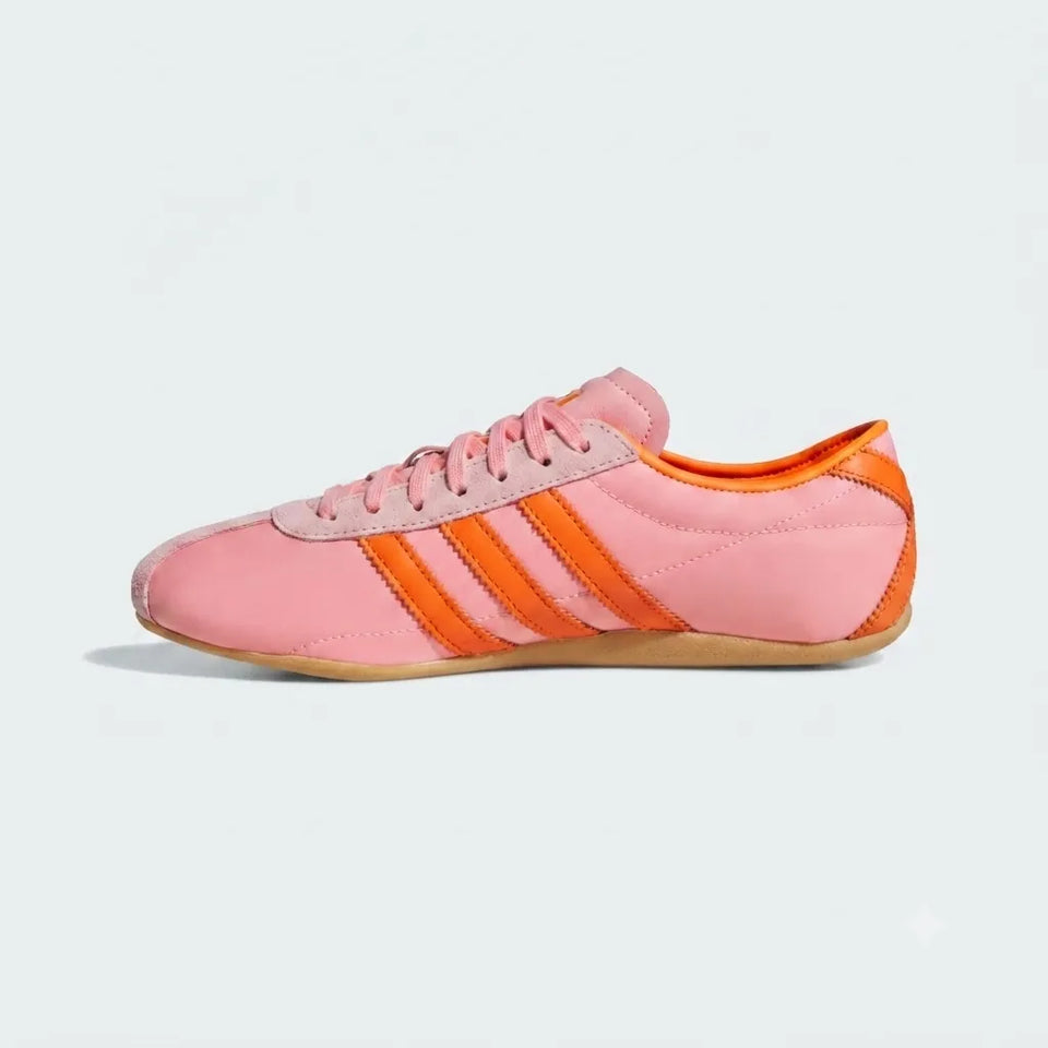 A pink low-top sneaker with three orange stripes on the side, pink laces, and a gum-colored sole, shown in profile against a light gray background.