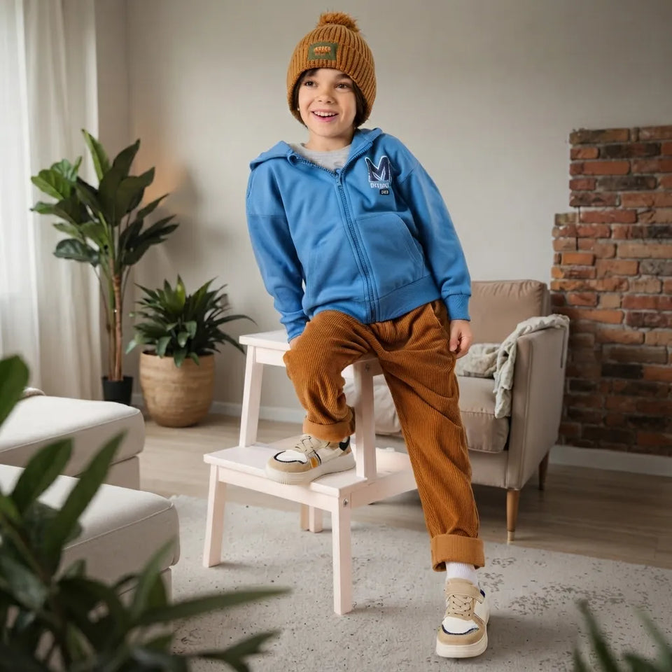 A child wearing a blue zip-up hoodie, rust-brown corduroy pants, a mustard knit beanie, and cream sneakers stands on a small wooden stool in a modern living room setting.