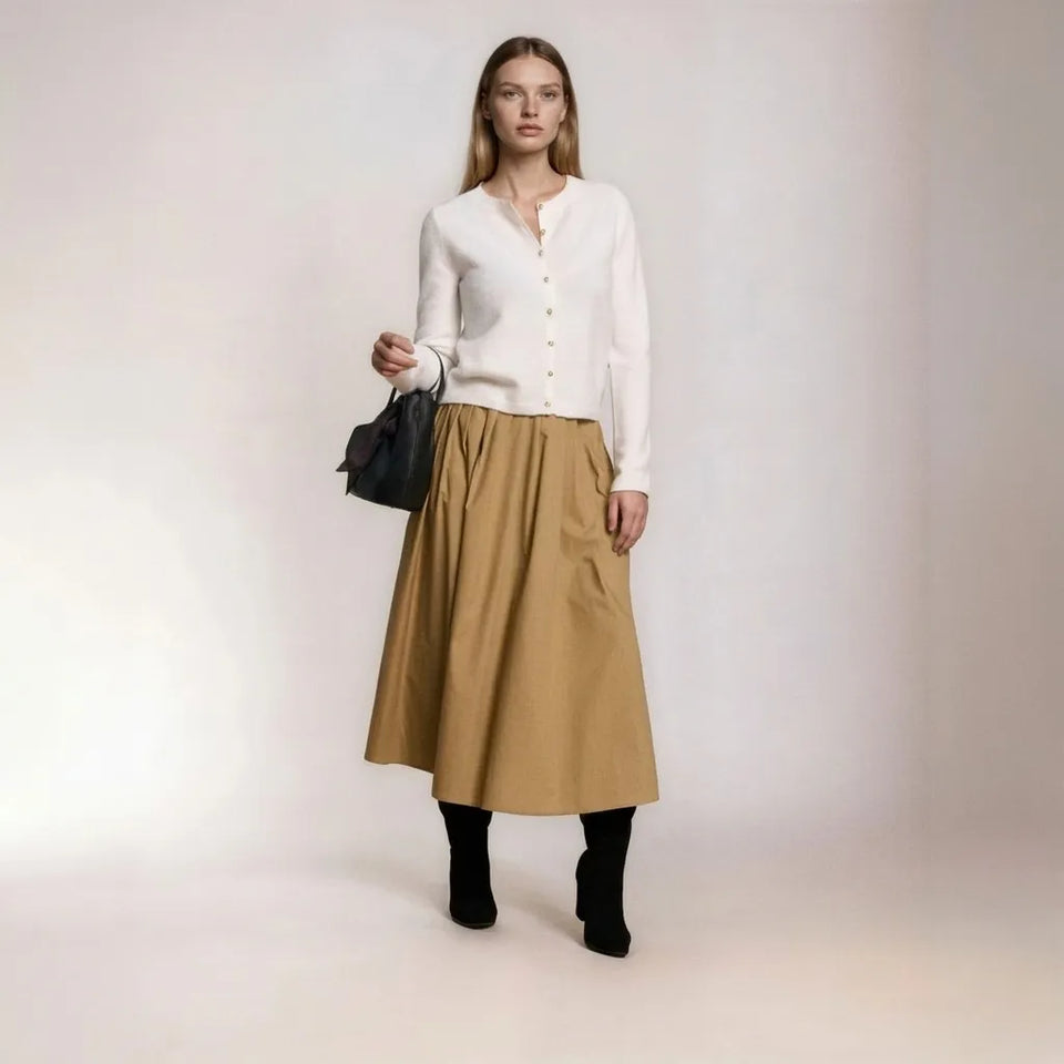 A woman stands against a plain light studio background wearing a white long-sleeve button-front cardigan, a knee-length tan skirt with side pockets, black ankle socks, and black ankle boots, while carrying a small black handbag.
