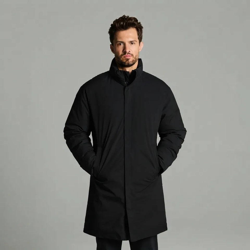 A man wearing a black mid-length puffer coat with a high stand-up collar, standing against a solid gray studio background.