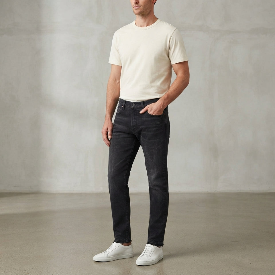 A man wearing dark charcoal slim-fit jeans, a cream t-shirt, and white sneakers, standing in a studio with a textured concrete wall and floor.